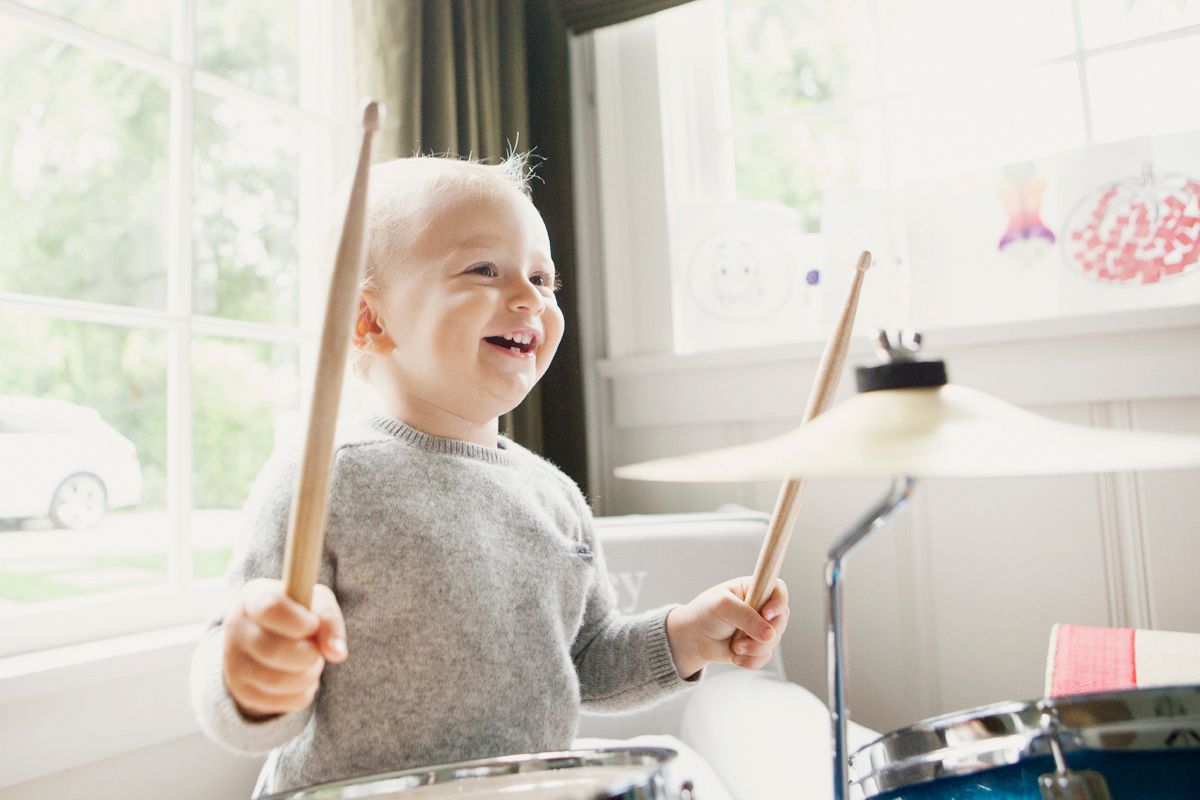 Happy toddler boy smiling while playing drums with drumsticks in a bright home living room.