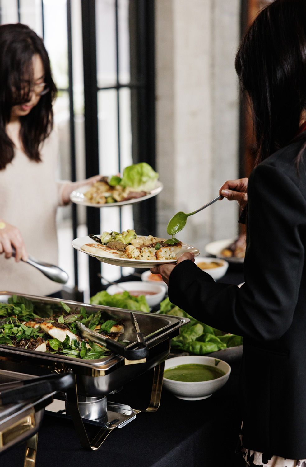 Two women serving themselves food at an elegant catered buffet with fresh salads and green garnishes.