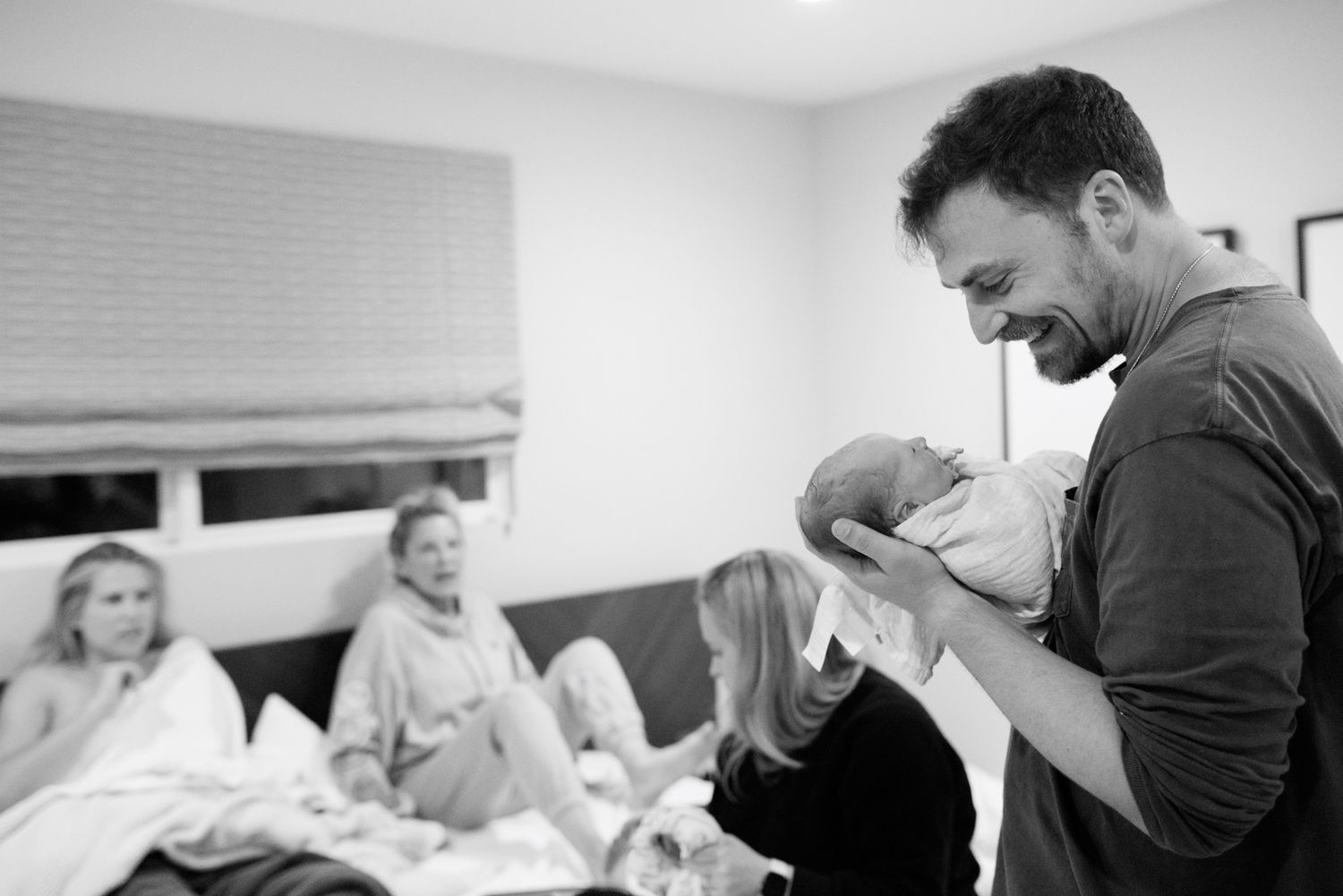 Smiling man cradles newborn baby while family relaxes on bed in background, candid black and white photo.