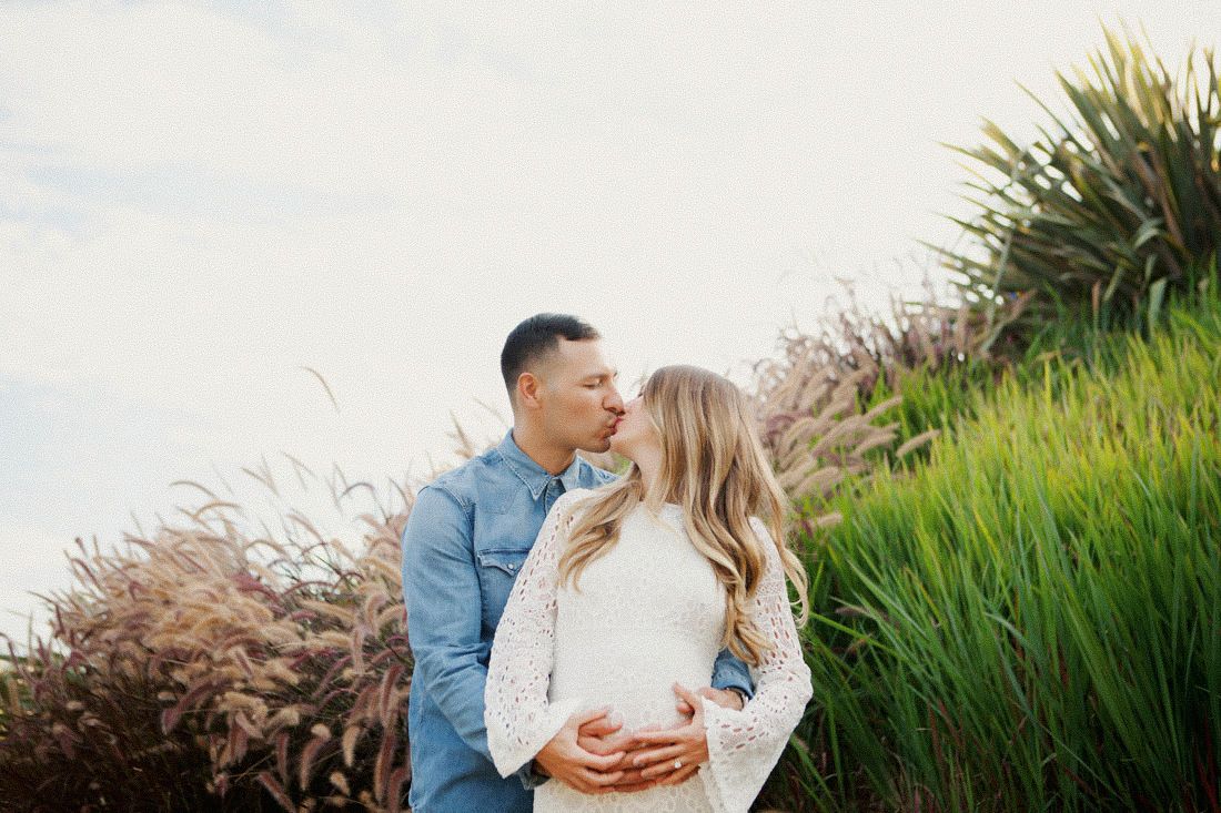 A couple shares a tender kiss outdoors, surrounded by lush green grasses and plants in a romantic engagement photo.