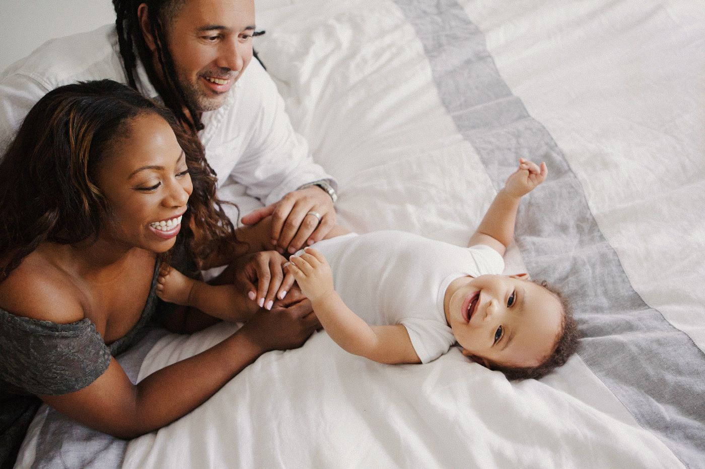 Happy family with parents smiling at their baby lying on a white bed in a warm, joyful moment.