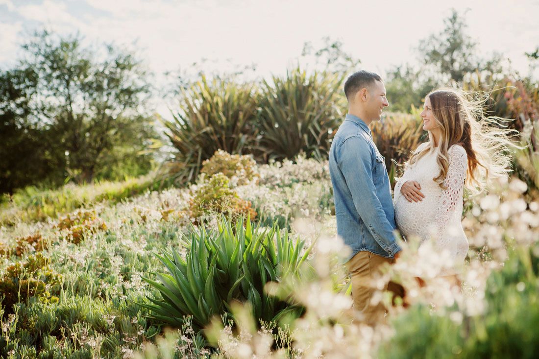 A couple stands together in a sunlit wildflower garden, sharing a tender moment surrounded by lush greenery.