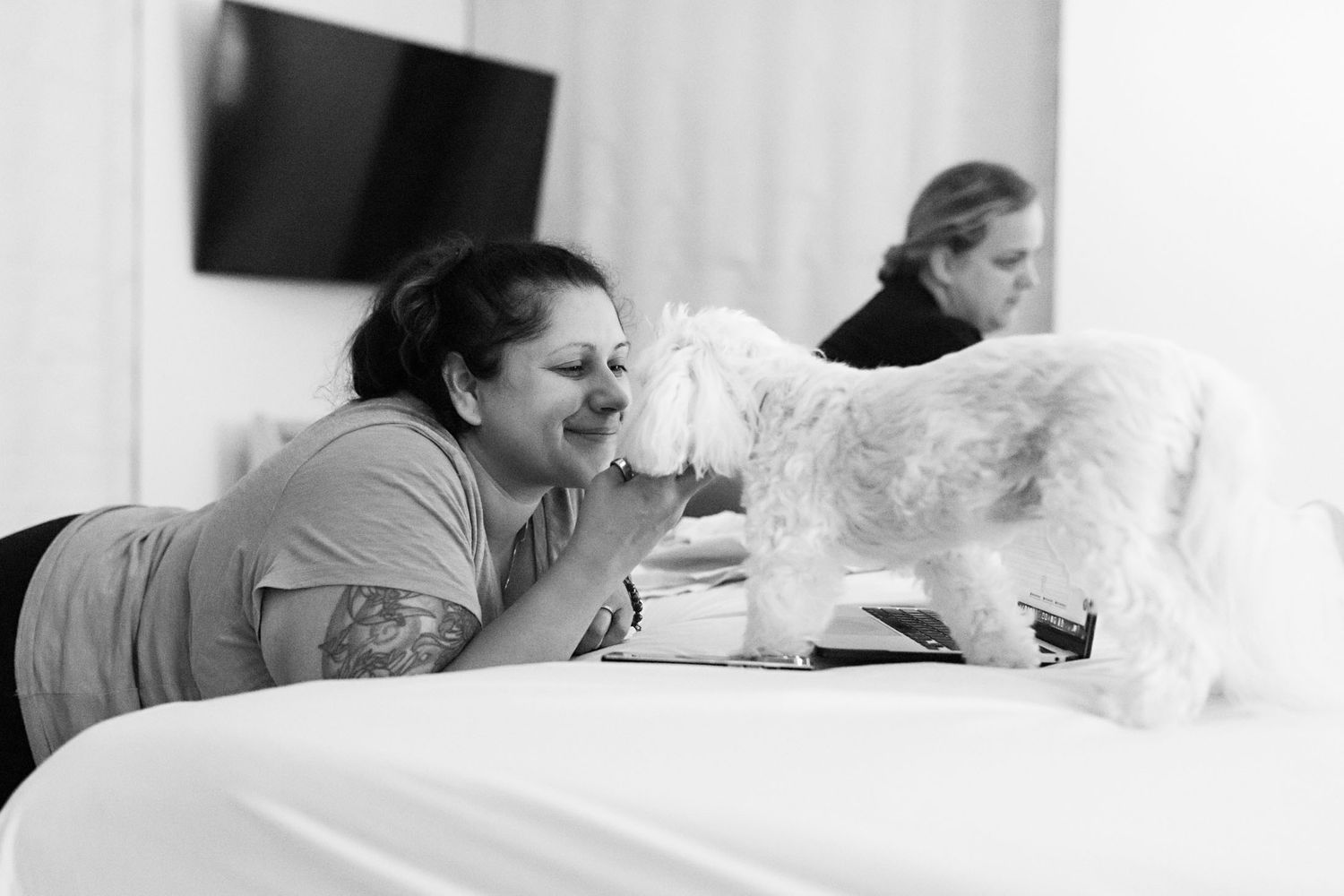 Black and white photo of a woman smiling as a fluffy white dog licks her face on a bed.