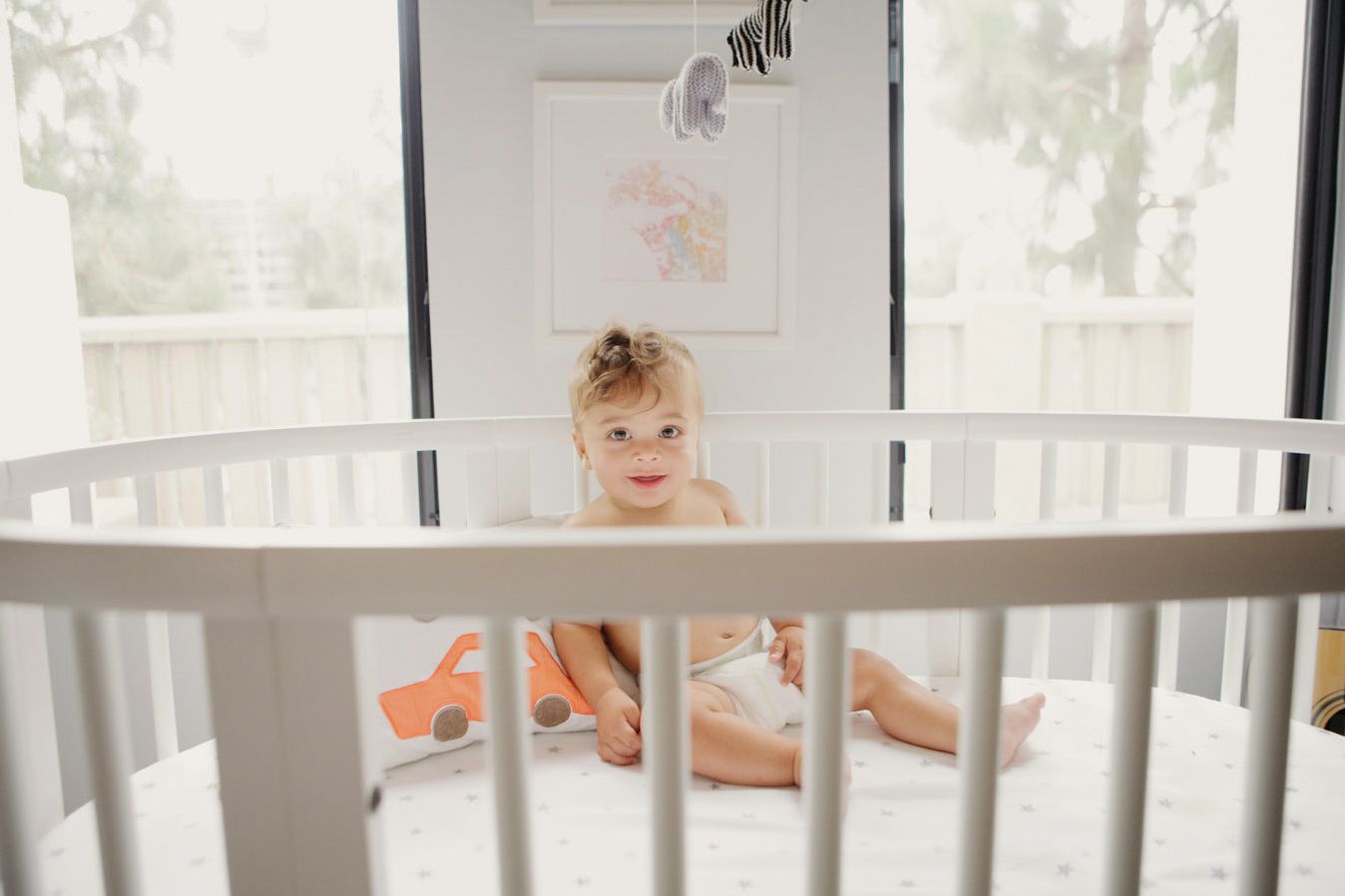 Smiling baby sitting in a white oval crib in a bright nursery room with soft decor.