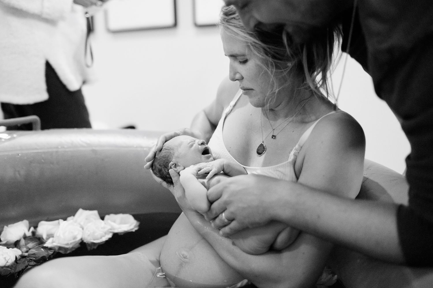 Black and white image of a mother holding her newborn in a water birth pool surrounded by floating white flowers.