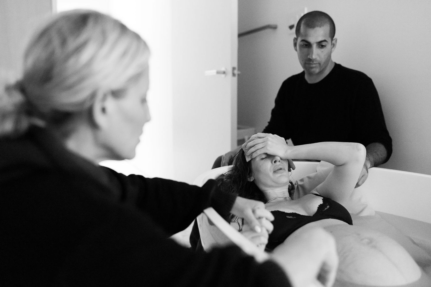 Black and white photo of a pregnant woman resting while a man watches and a midwife assists during home labor.