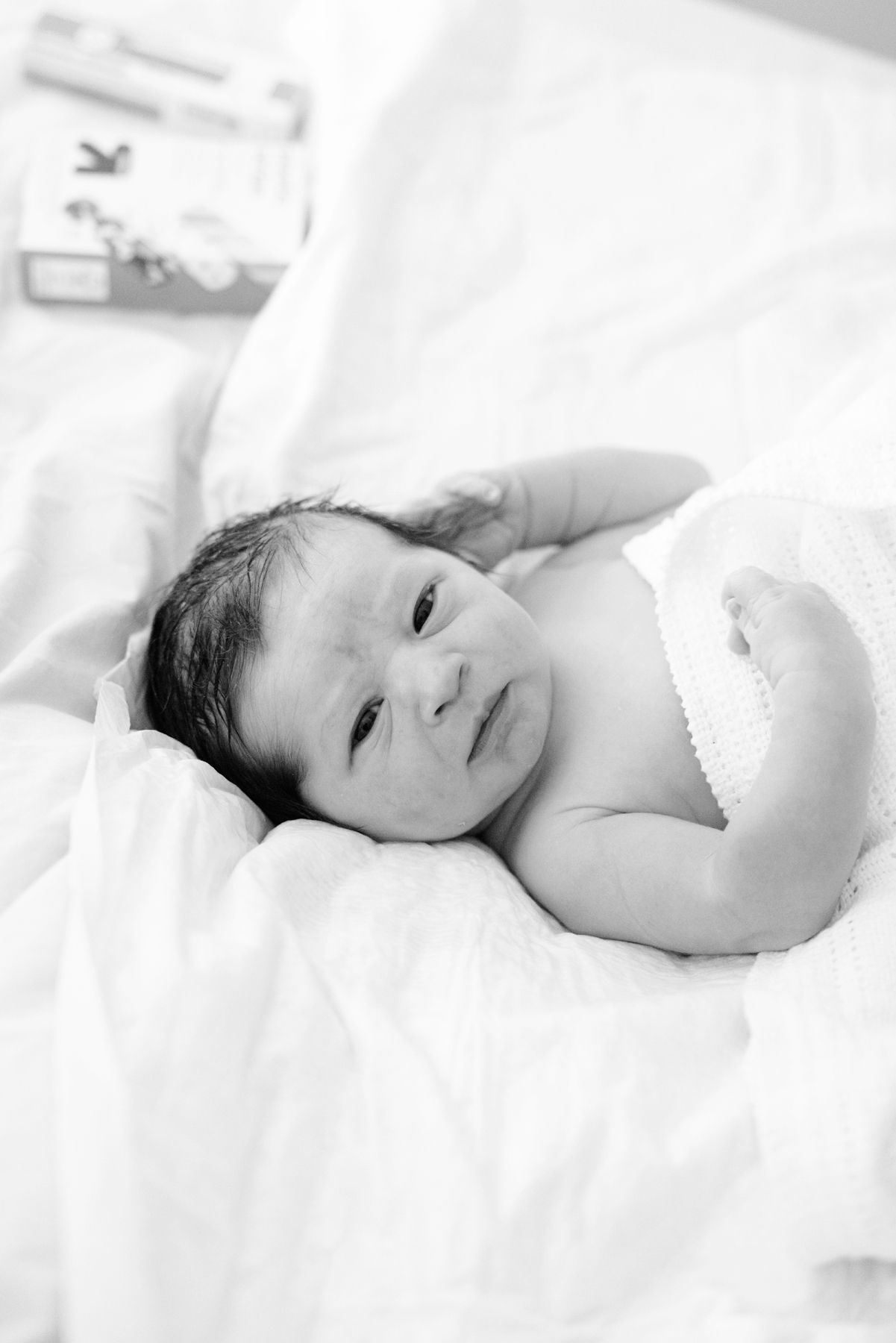 Peaceful newborn baby with dark hair lying on white blankets, serene black and white portrait.