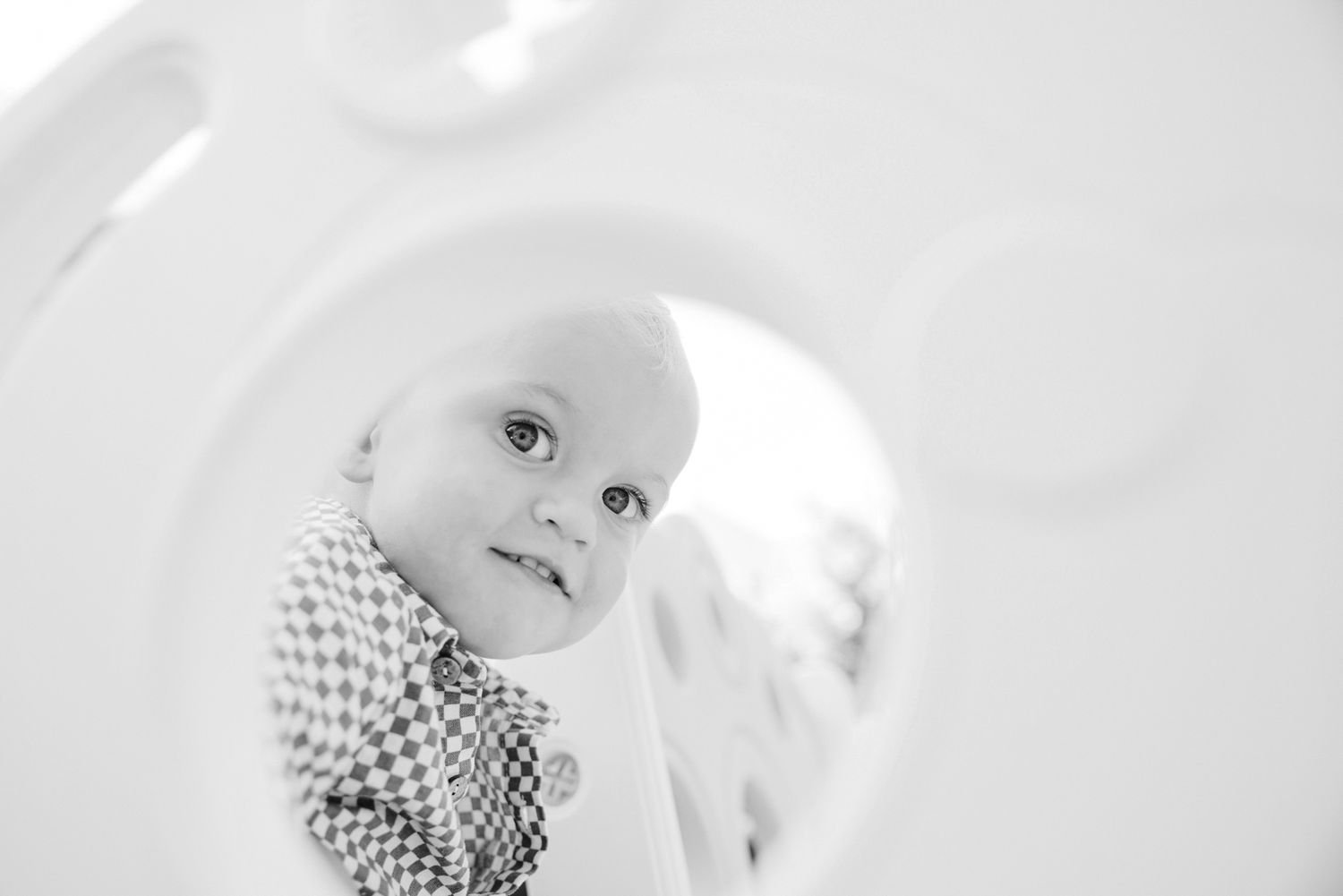 Black and white photo of a smiling baby peeking through a circular opening, wearing a checkered outfit.