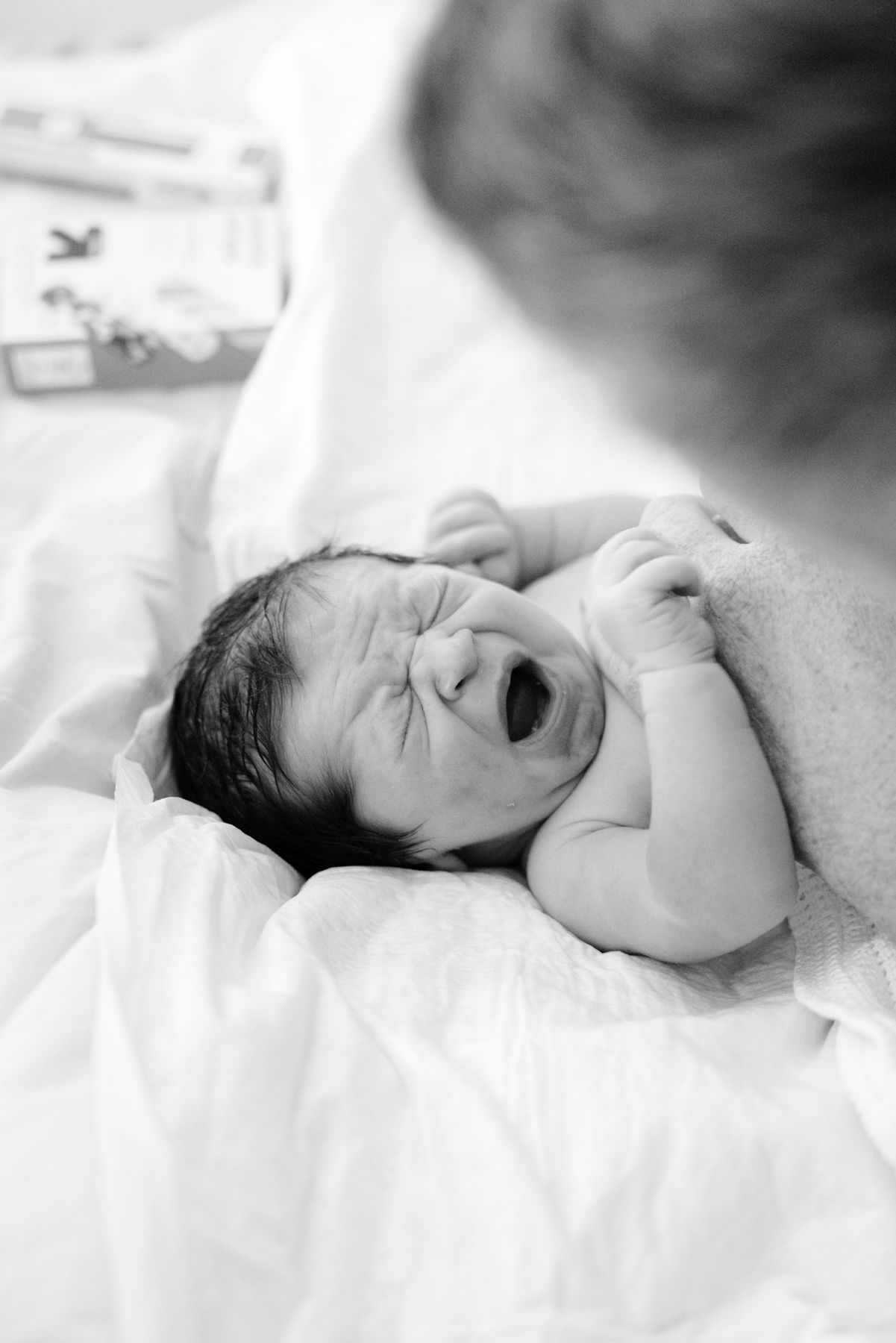 Crying newborn baby lying on white blankets in hospital, black and white birth photography.