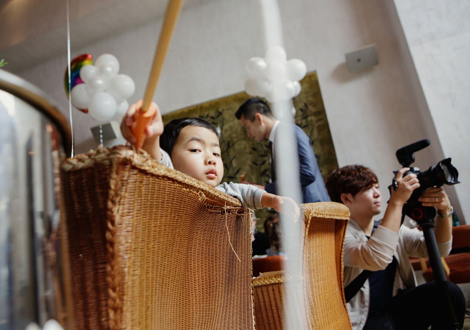 Young Asian child peers over wicker basket at party venue with balloons and photographer nearby.