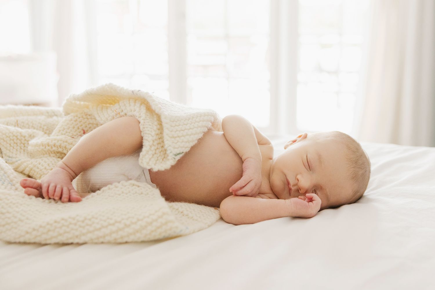 Peaceful newborn baby sleeping on white bed covered with soft cream knit blanket in bright airy room.