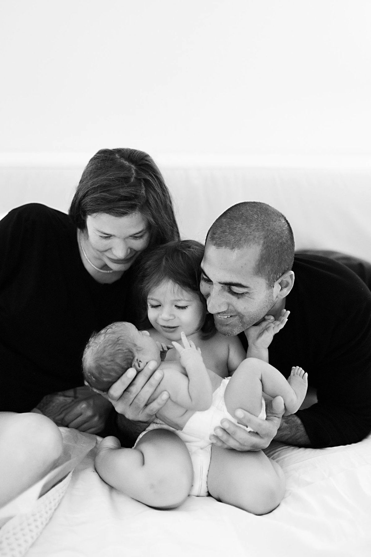 Black and white family photo of parents and toddler lovingly holding a newborn baby on a white bed.