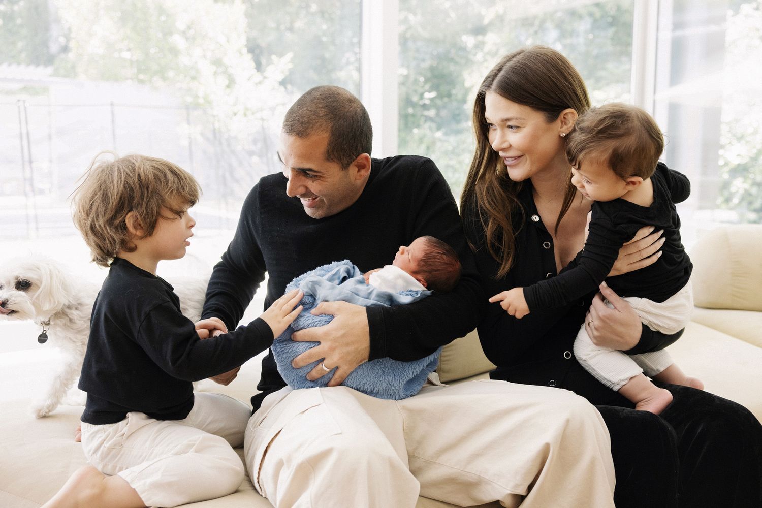 Happy family of five sitting on a couch, parents holding newborn baby and toddler, with older child beside them.