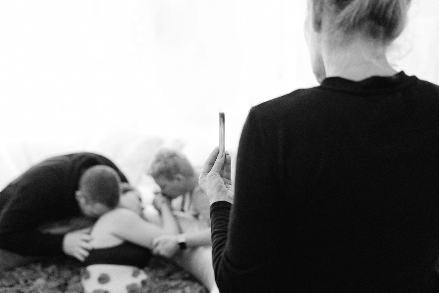 Black and white photo of woman holding phone while two people embrace on bed in blurred background.