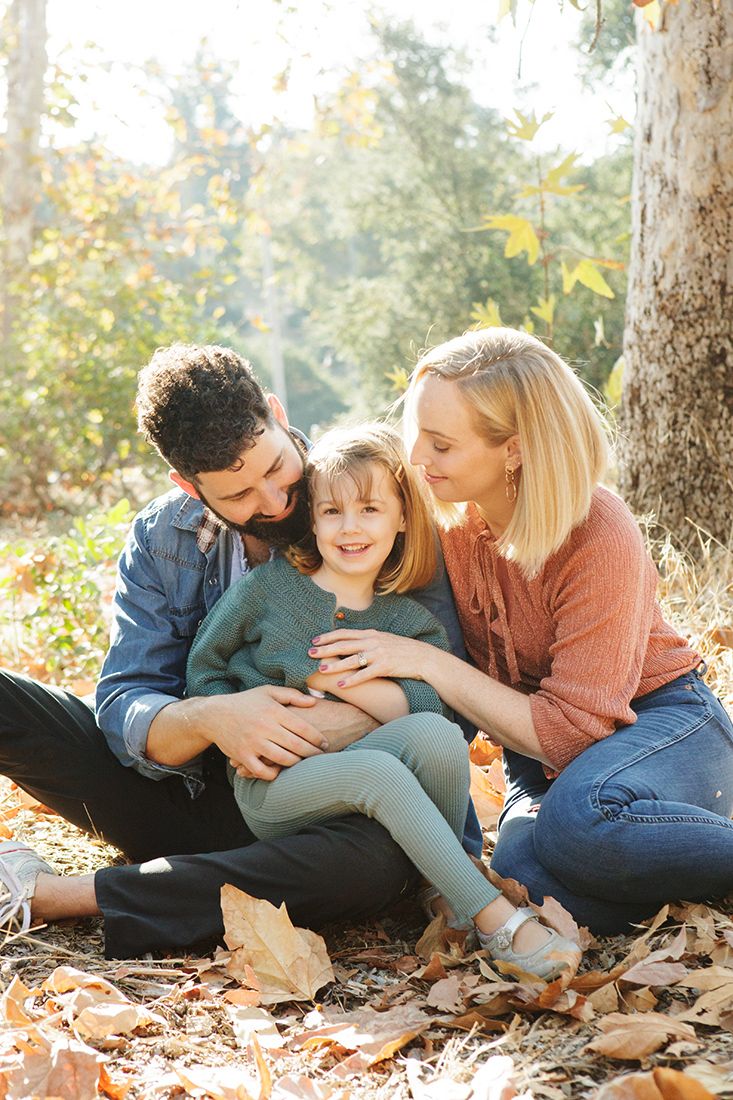 Happy family of three sitting together among autumn leaves in a sunny fall forest setting.