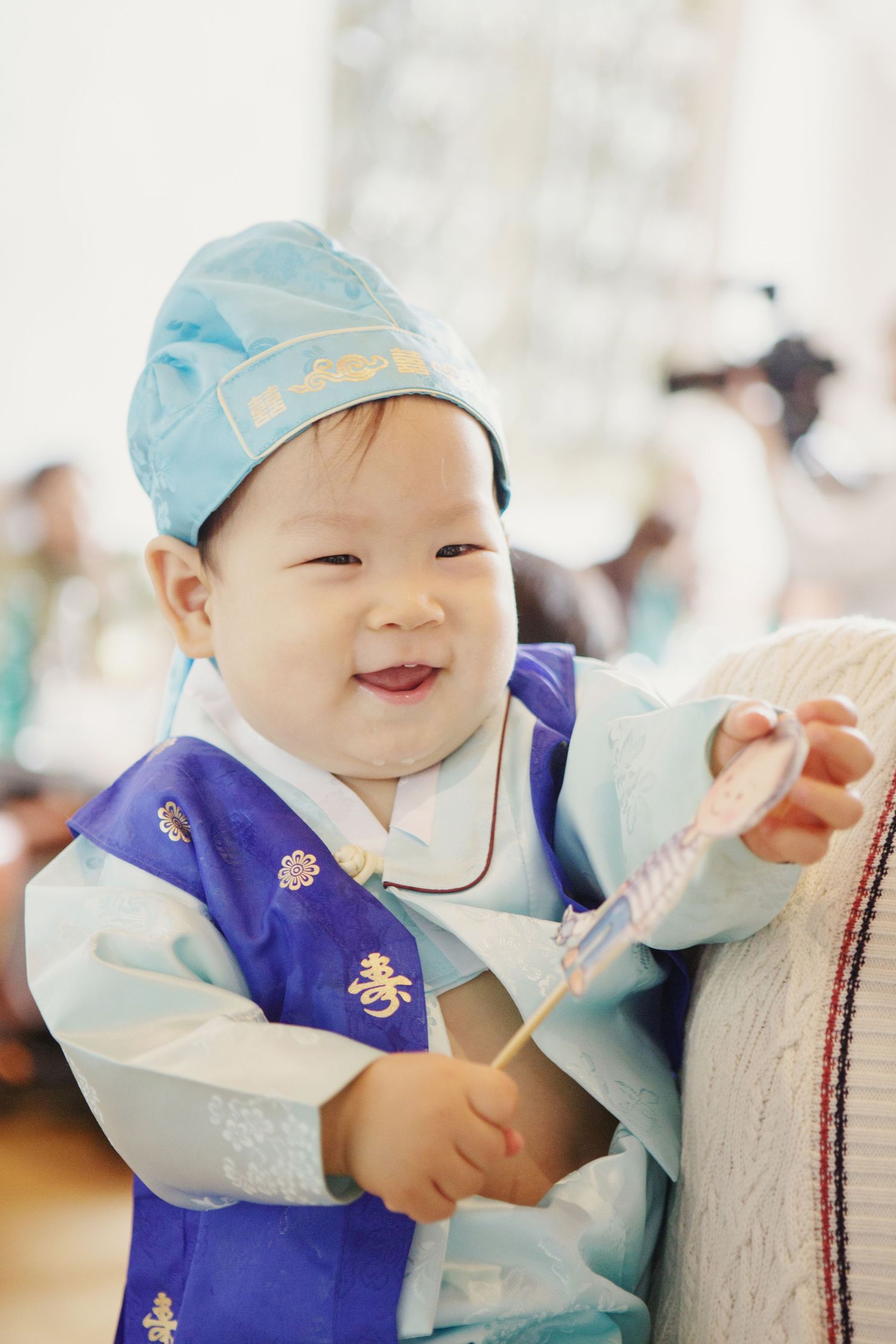 Happy Asian baby in blue hanbok and hat smiling during traditional Korean dol birthday celebration.