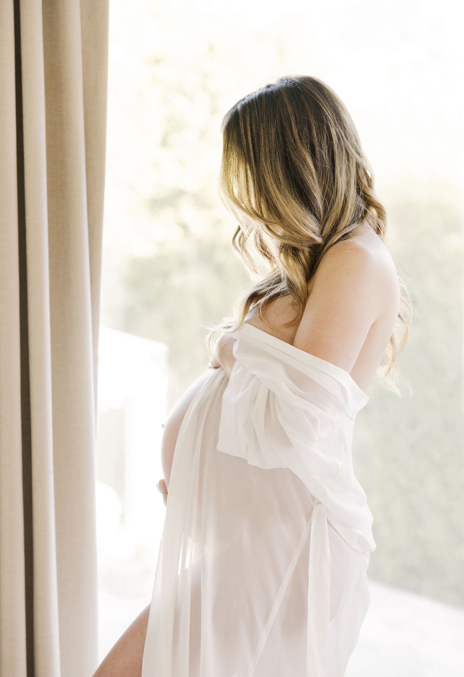 Pregnant woman in sheer white robe standing by window, showcasing beautiful maternity photography in soft natural light.