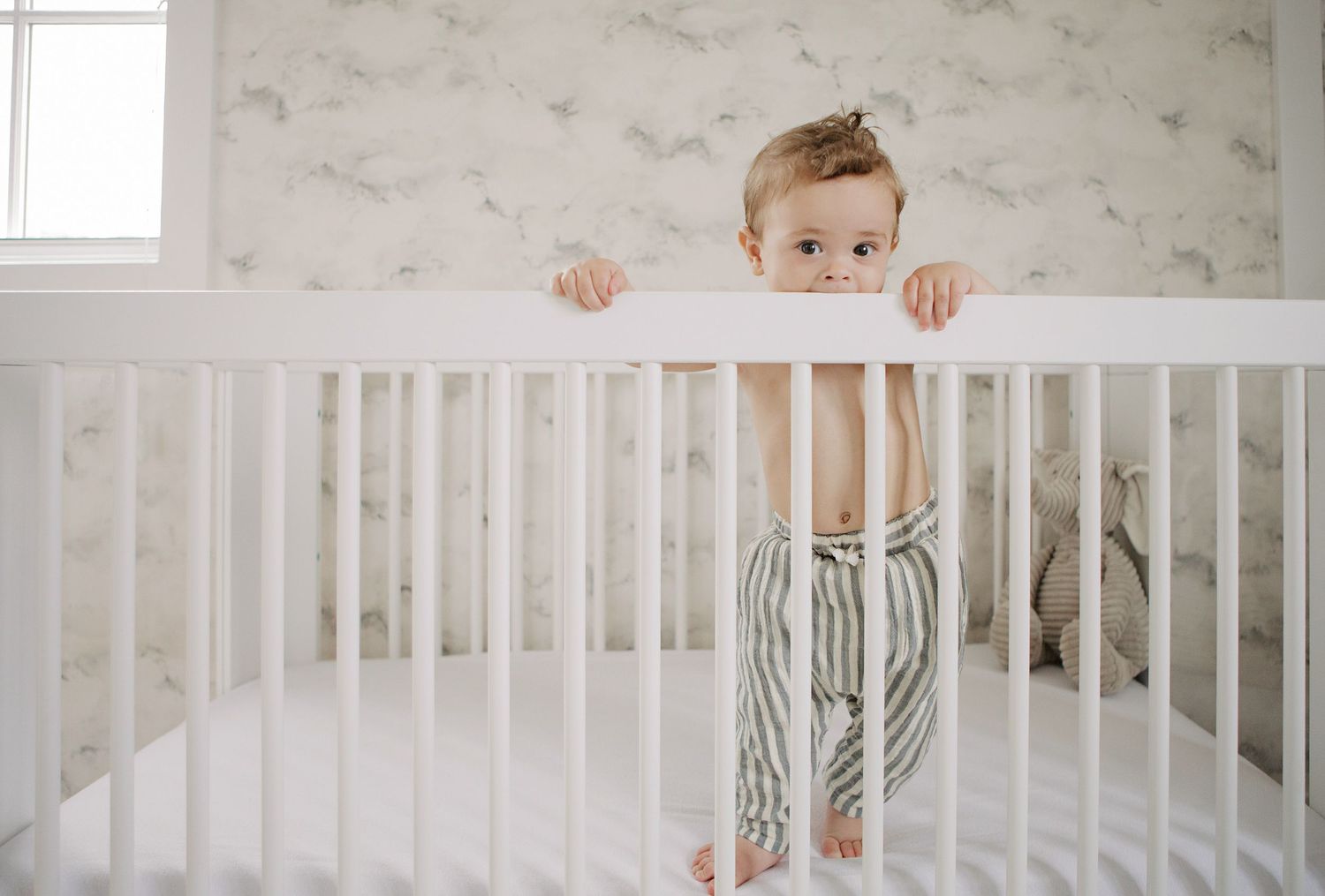 Smiling baby standing in a white crib wearing striped pants in a soft nursery with floral wallpaper.