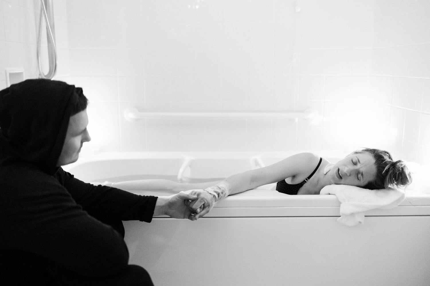 Black and white photo of a person holding hands with a woman resting in a bathtub.