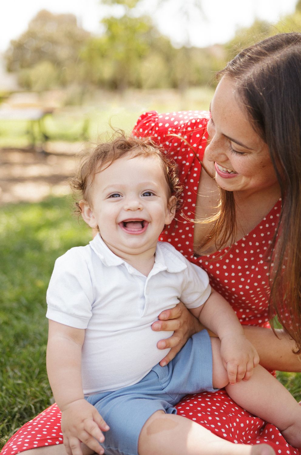 A joyful baby laughing outdoors while sitting on a smiling woman in a red polka dot dress on green grass.