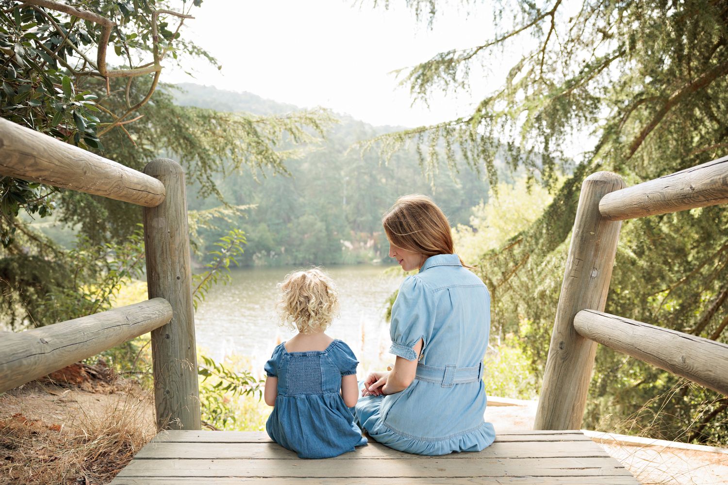 Mother and young daughter sitting together on wooden deck overlooking a serene lake surrounded by lush green trees.