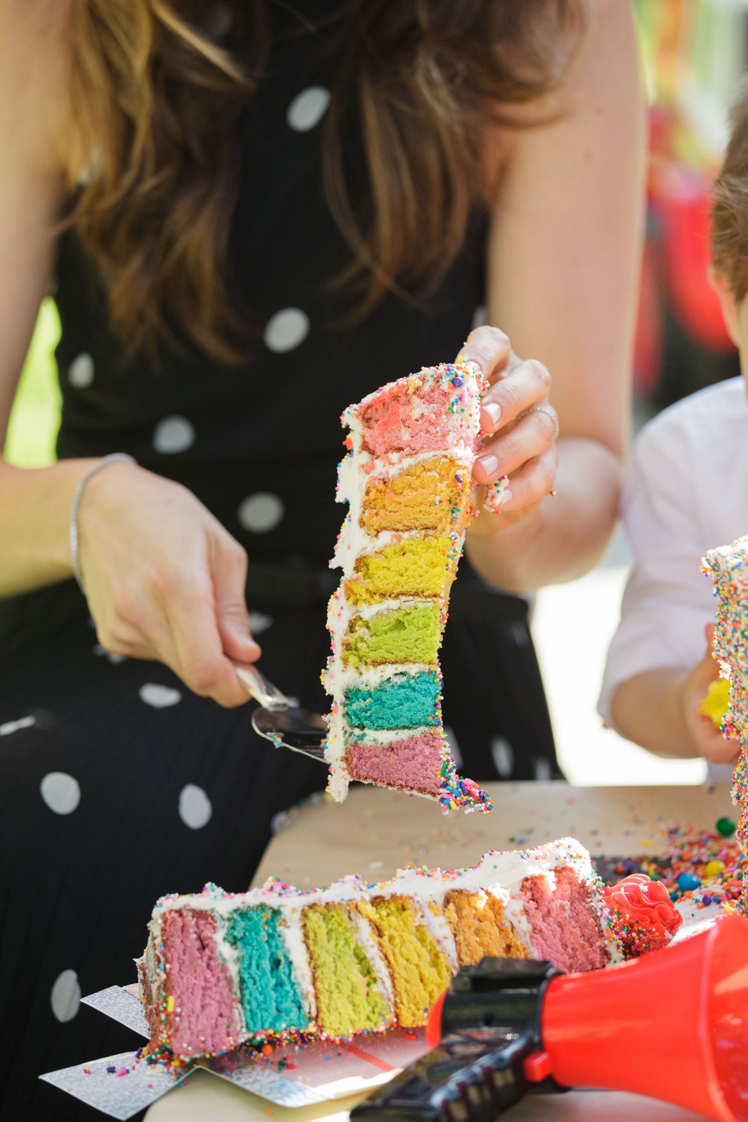 A colorful rainbow layer cake slice being lifted, showing vibrant multicolored layers with white frosting and sprinkles.