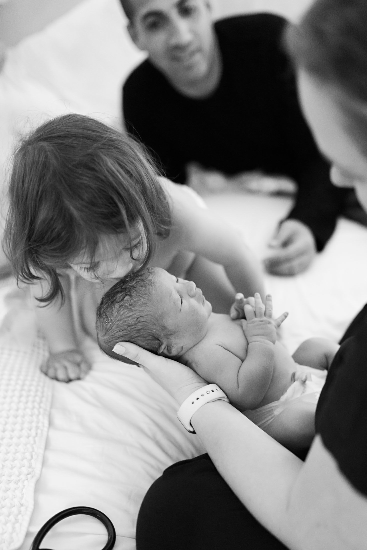 A toddler leans in to kiss a newborn baby held by a mother as father watches in a black and white photo.