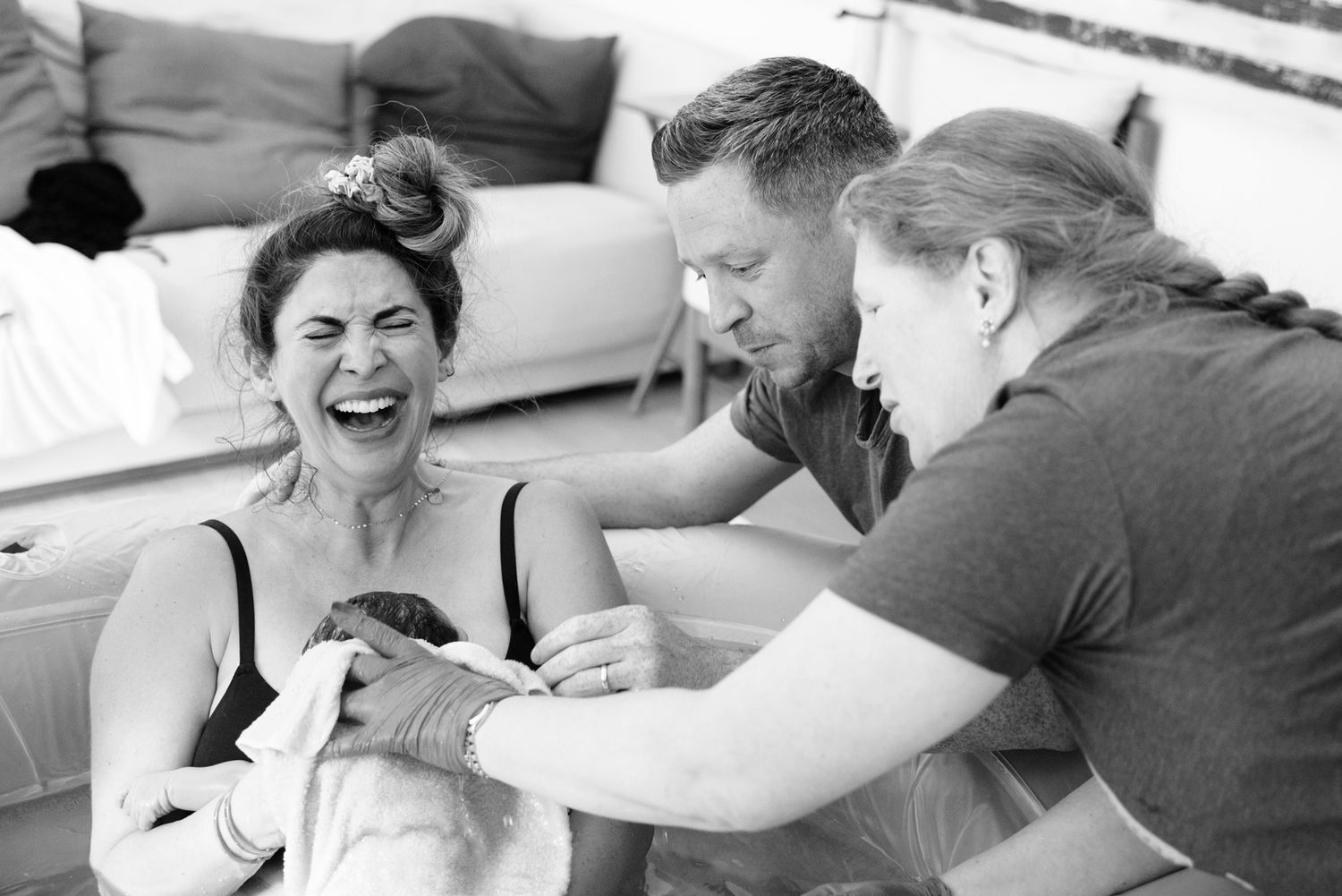 New mother laughs joyfully while holding newborn assisted by partner and midwife after home birth.