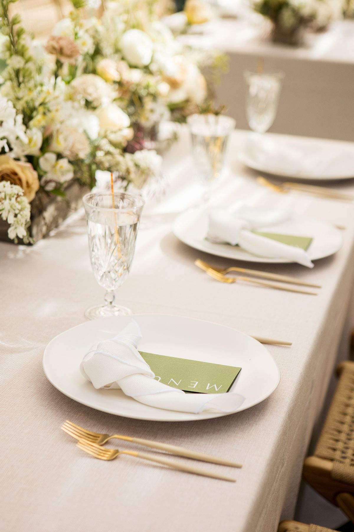 Elegant wedding table setting with gold cutlery, white plates, green menu cards, and crystal glassware on a neutral linen tablecloth.