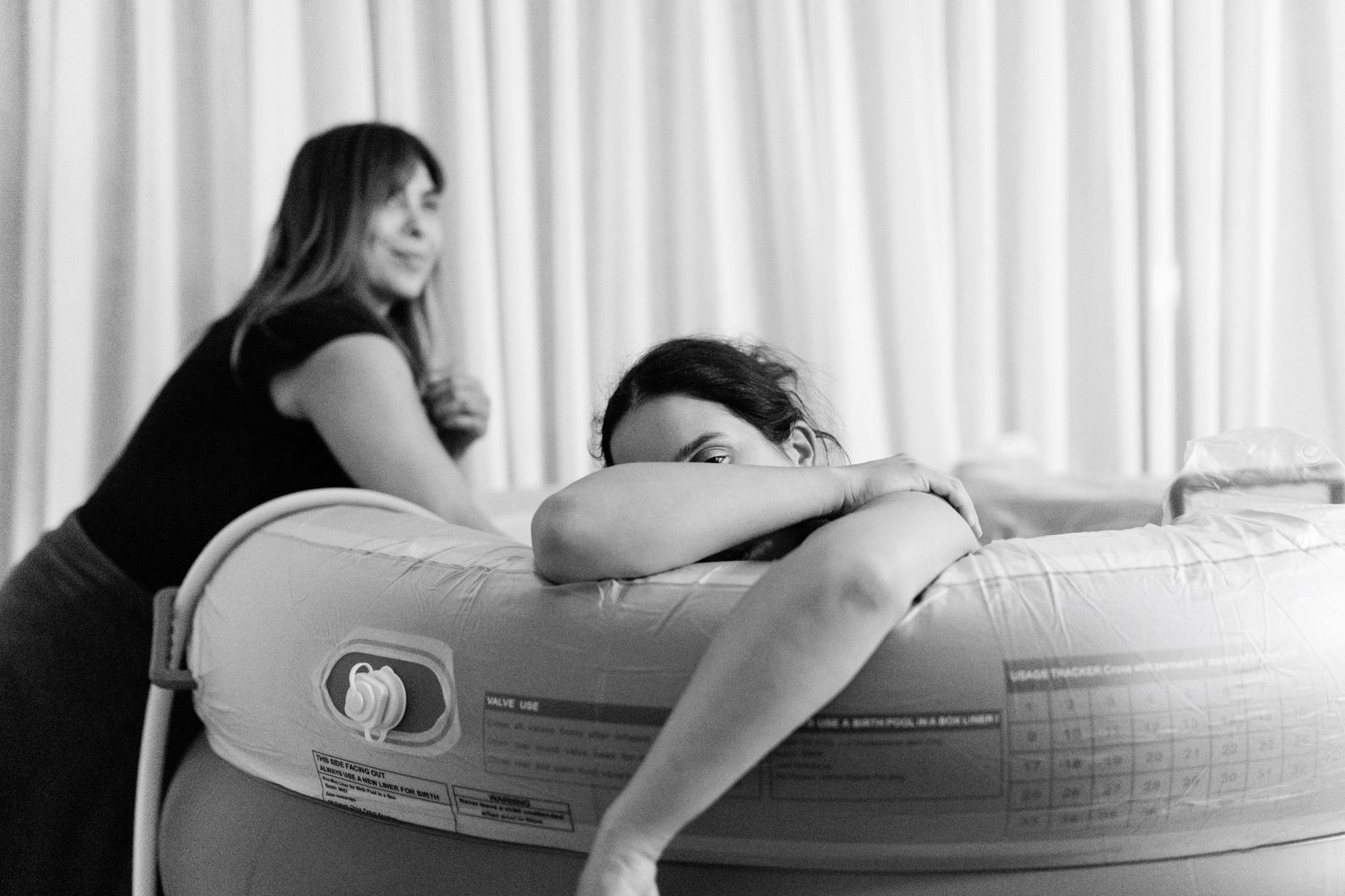 Black and white photo of a woman leaning on an inflatable birth pool during labor, supported by a doula.