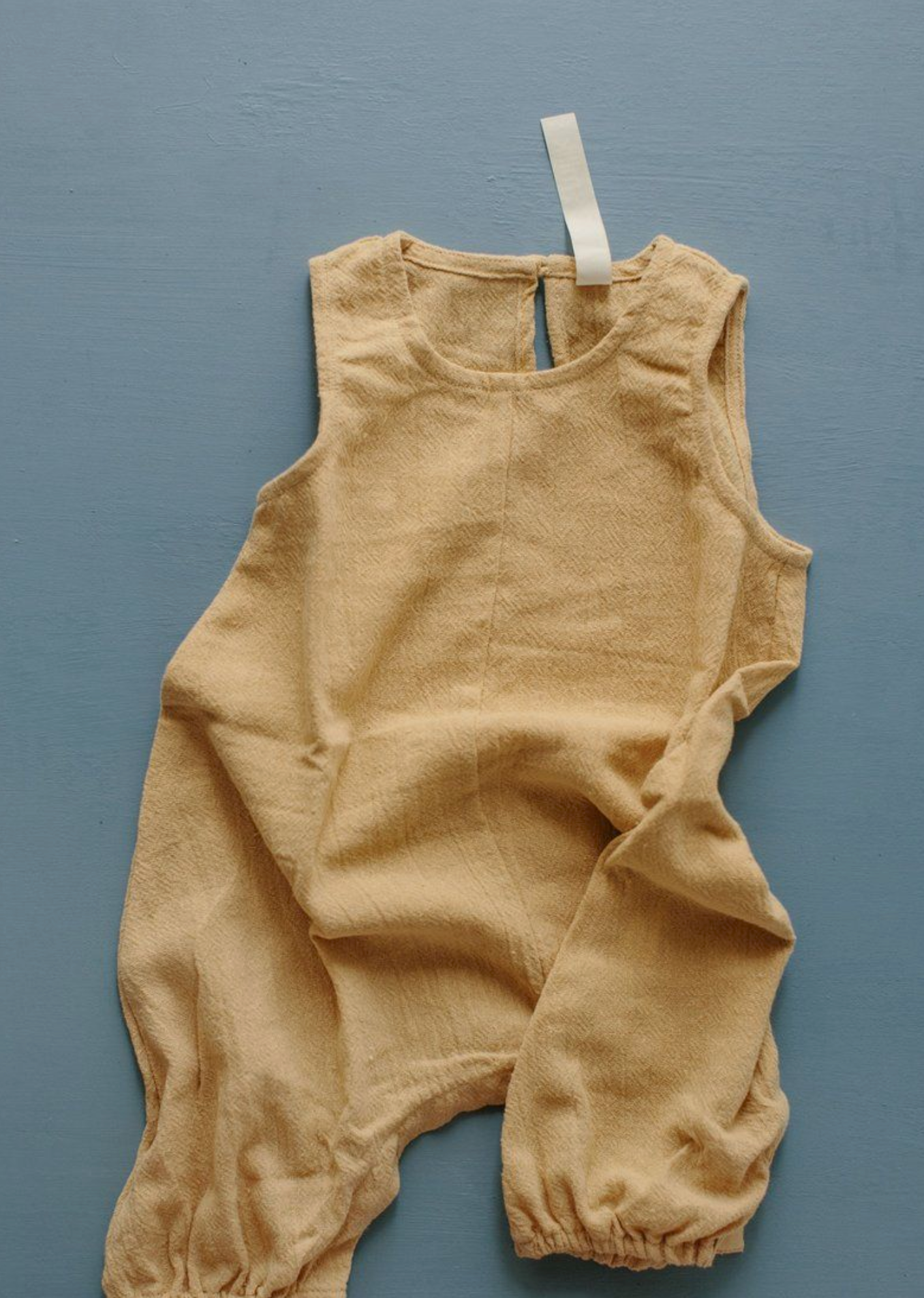 Tan linen sleeveless romper with elastic cuffs and back button closure, laid flat on blue background.