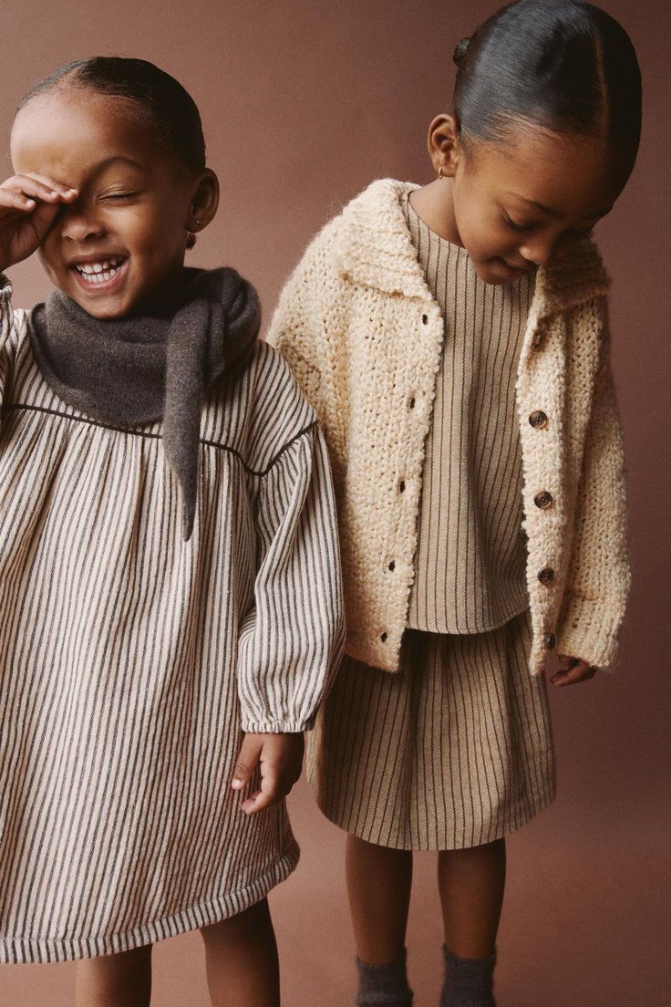 Two young girls in neutral-toned children's fashion, one in striped dress with scarf, other in ribbed set with knit cardigan.
