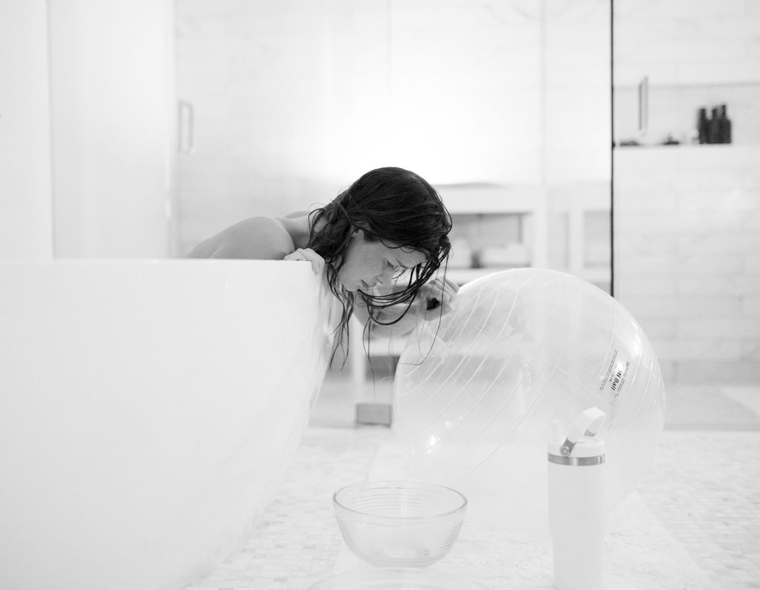 Black and white photo of a woman leaning over a bathtub with a glass bowl and lamp on a marble surface.
