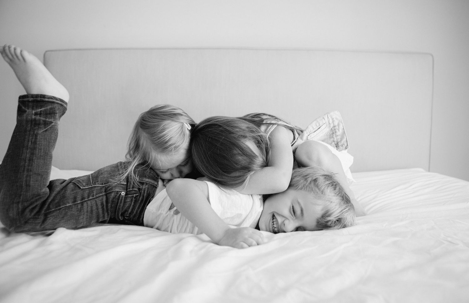 Three young children playfully cuddling and laughing together on a white bed in a black and white photo.