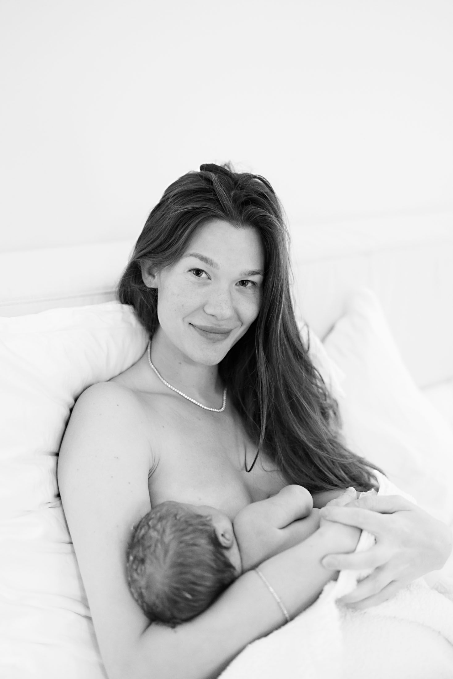 A smiling mother breastfeeds her newborn baby while resting against white pillows in a black and white photo.