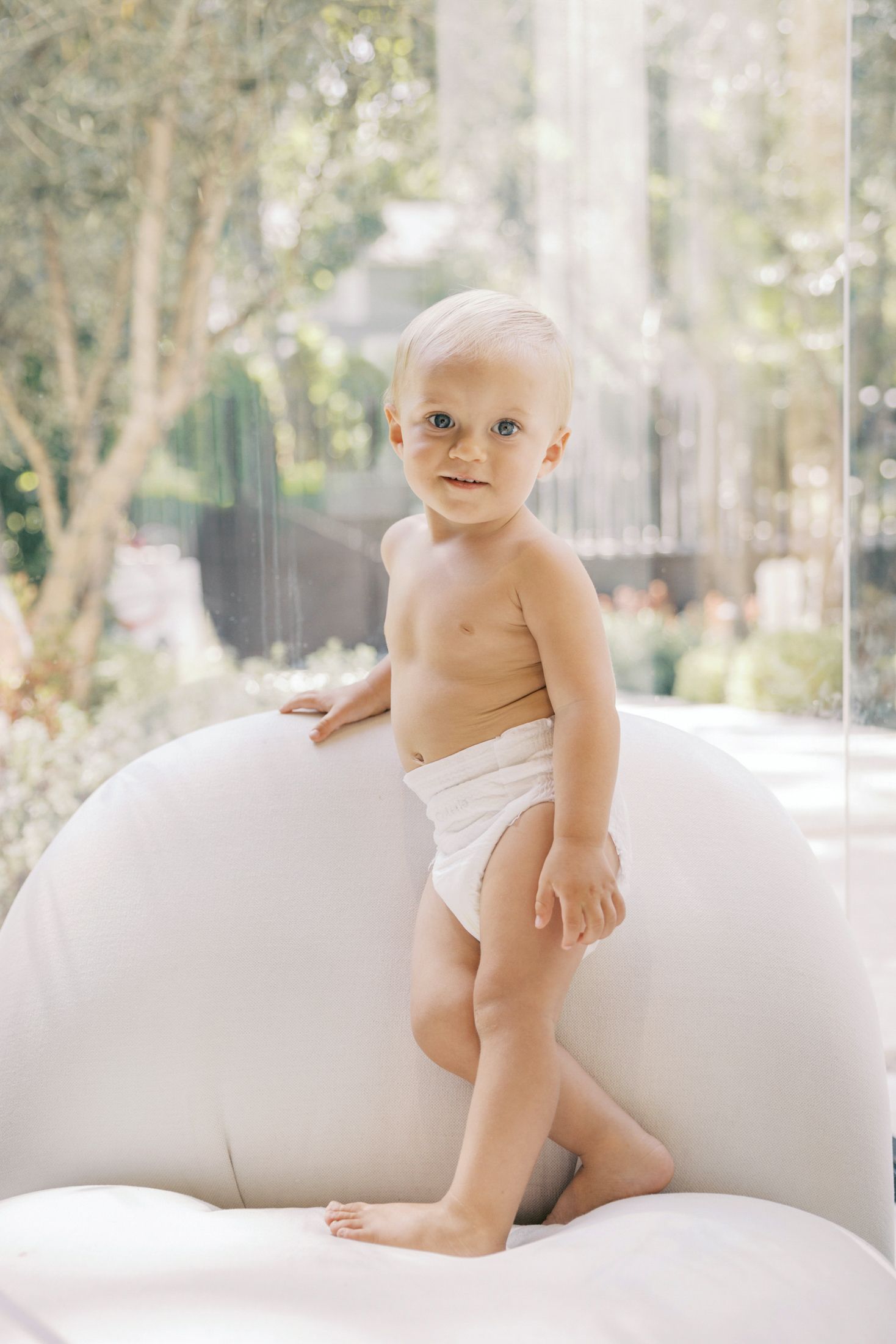 Adorable baby in white diaper standing on white chair with green garden background.