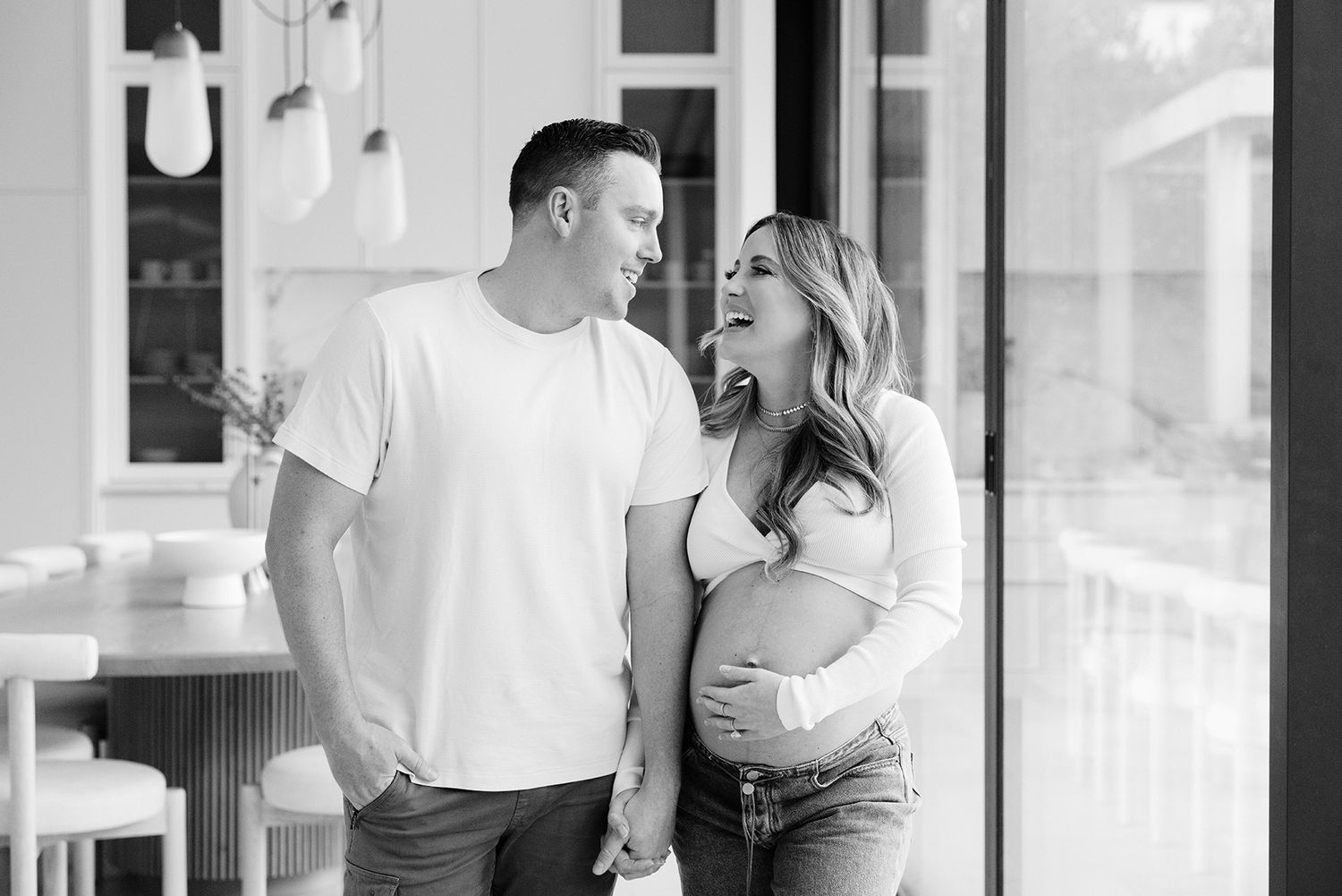 Black and white maternity photo of a couple smiling at each other, woman cradling baby bump, standing indoors.
