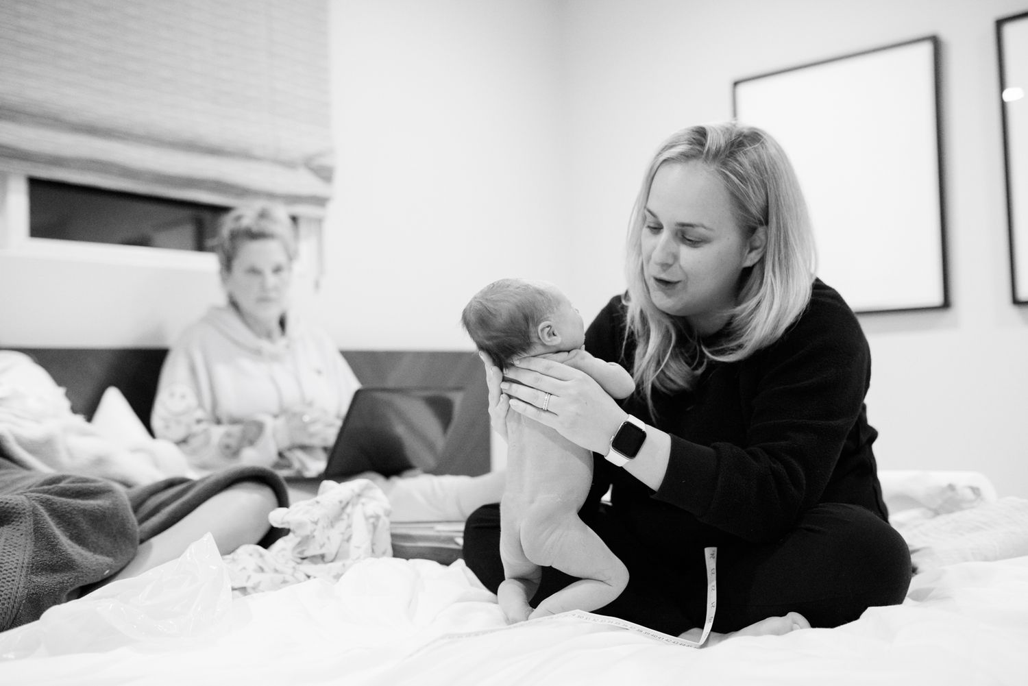 Woman in black holds newborn baby on white bed while another person rests in background, black and white photo.