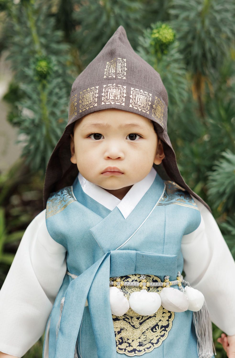 An adorable baby boy dressed in a traditional Korean hanbok and gat hat poses outdoors.