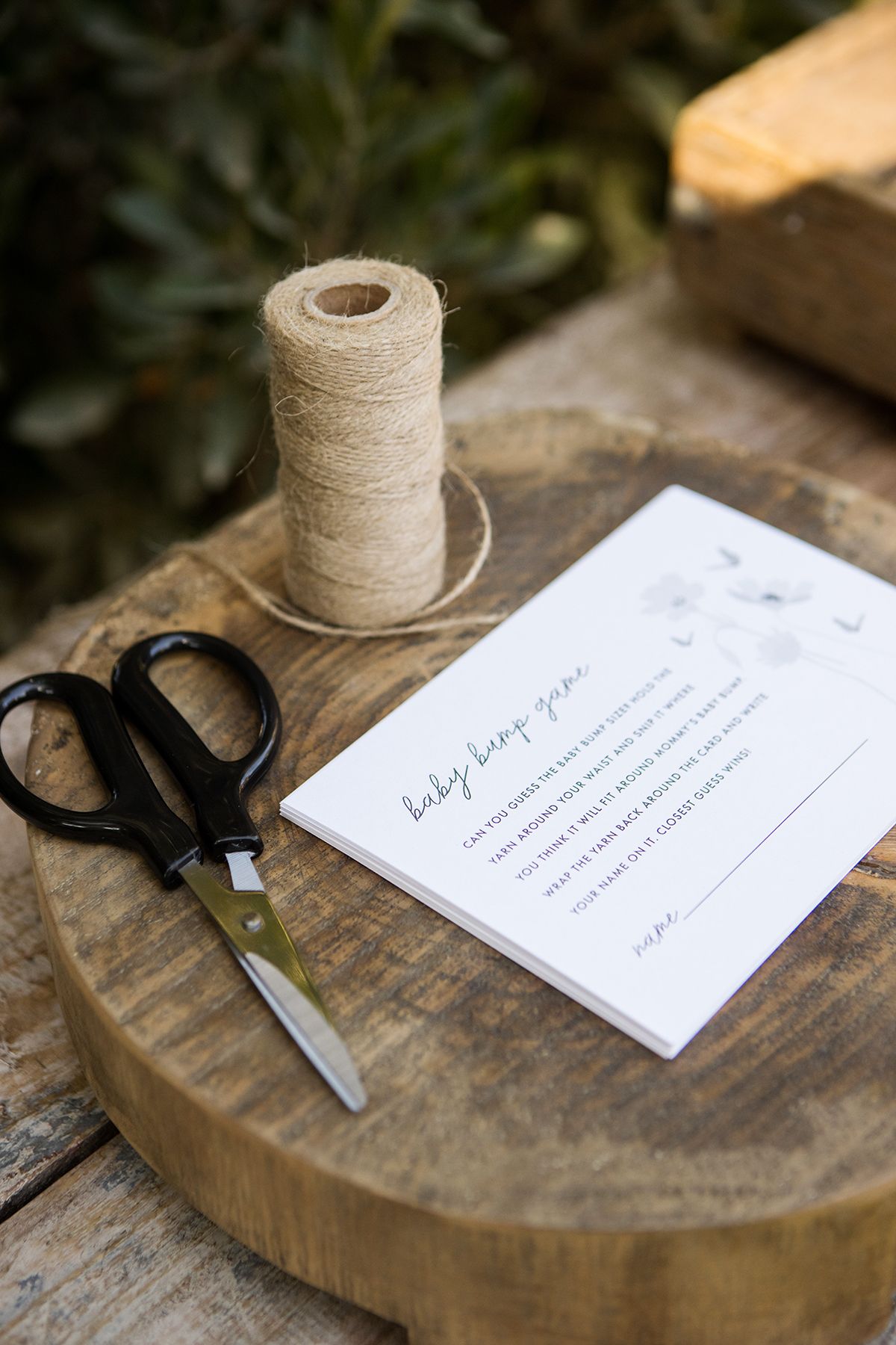 Baby bump game card with scissors and twine spool on a wooden slice at an outdoor baby shower.
