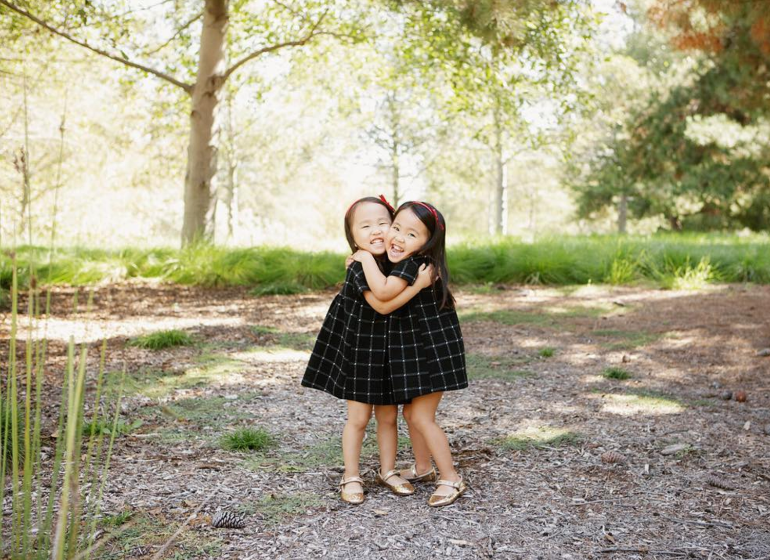 Two young girls in matching black dresses hugging outdoors in a sunlit park setting.
