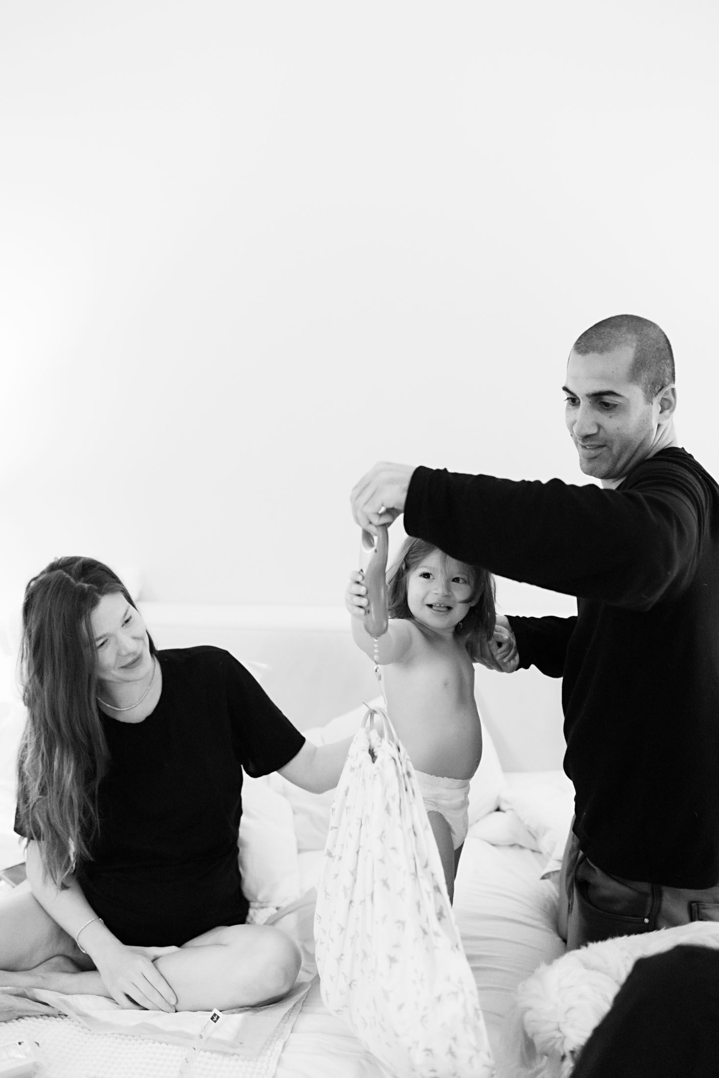 Parents smile as father helps toddler stand on a white bed beside a newborn in a black and white photo.