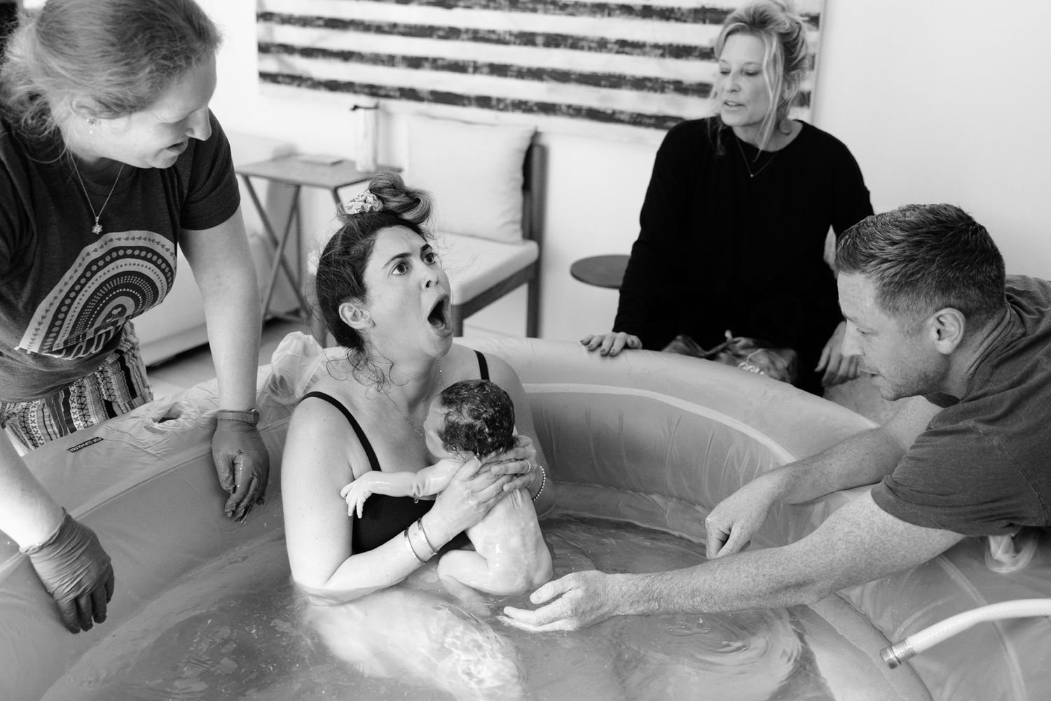 Mother holds newborn in water birth tub surrounded by supportive birth team and partner.