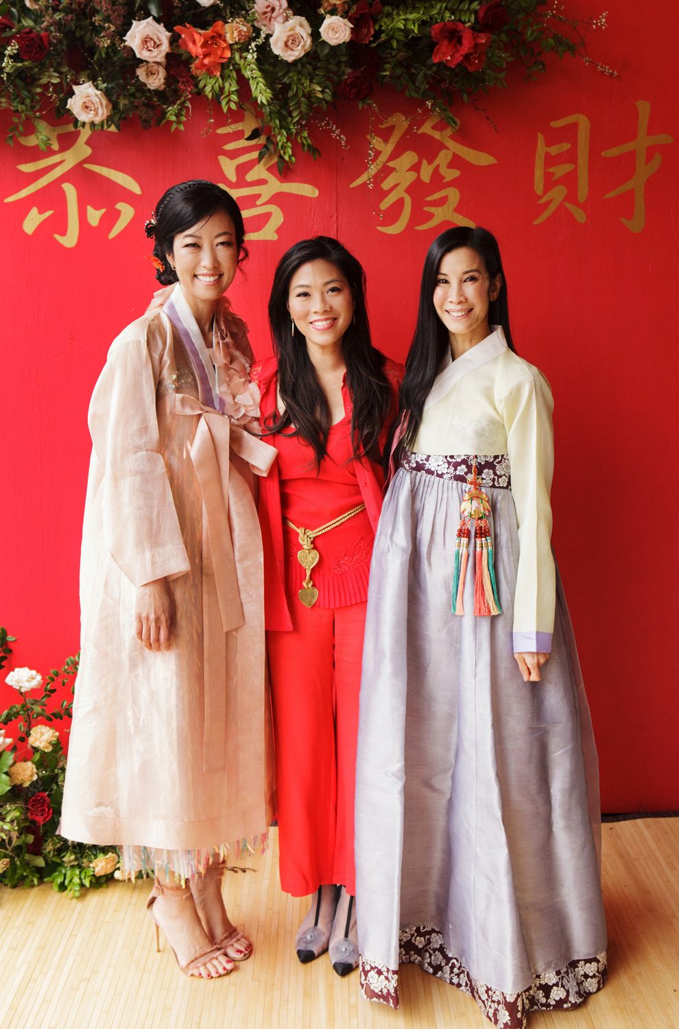 Three women posing in traditional Korean hanbok attire in front of a red backdrop with gold Chinese characters.