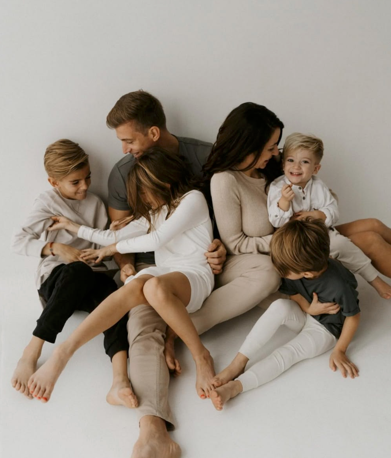 Happy family of six sitting together on white floor, parents with four young children in neutral tones.
