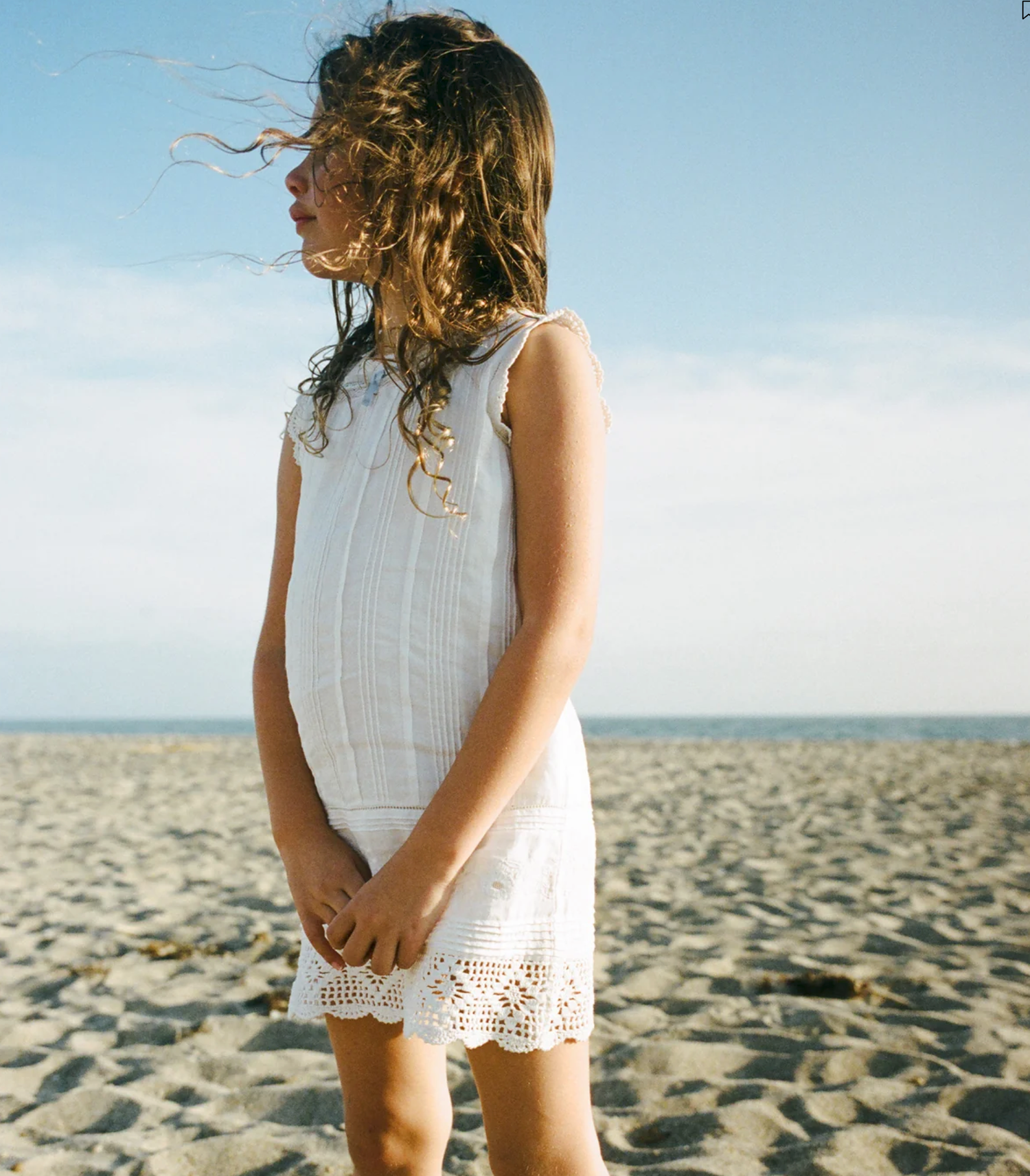 A young girl in a white lace dress stands on a sandy beach, her hair blowing in the wind on a sunny day.