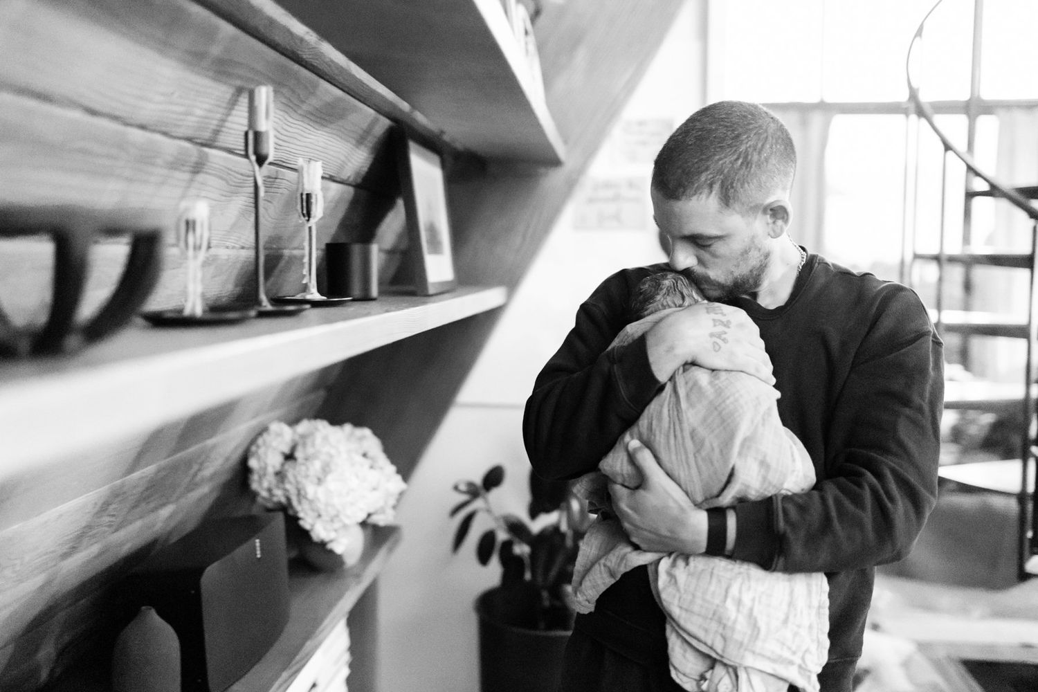 Black and white photo of a father tenderly kissing his newborn baby while cradling it in his arms indoors.