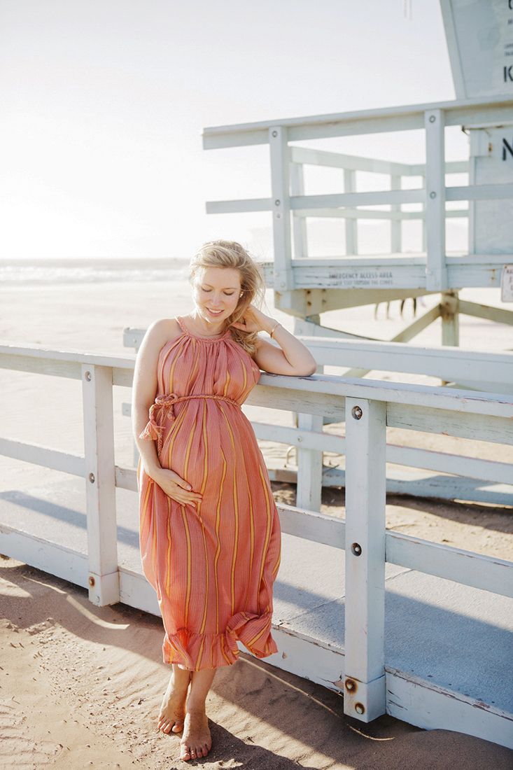 Pregnant woman in colorful striped maxi dress posing by a lifeguard tower on a sunny beach.