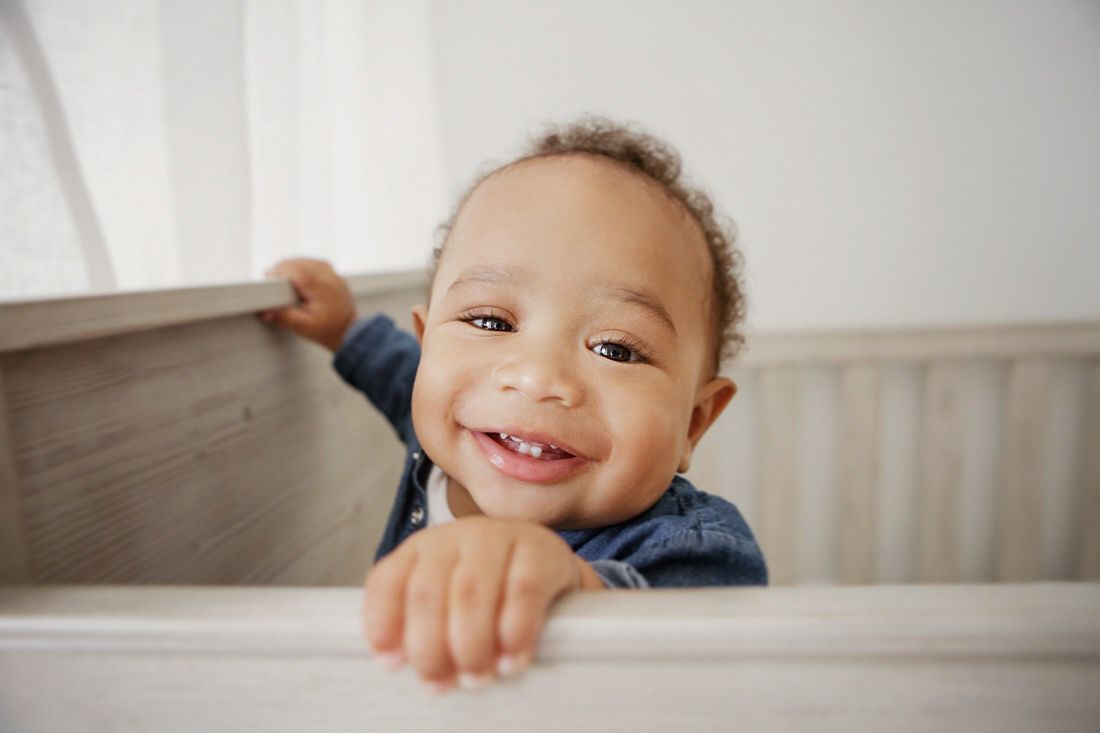 Smiling baby standing in a white wooden crib, wearing a denim shirt, with a joyful expression.