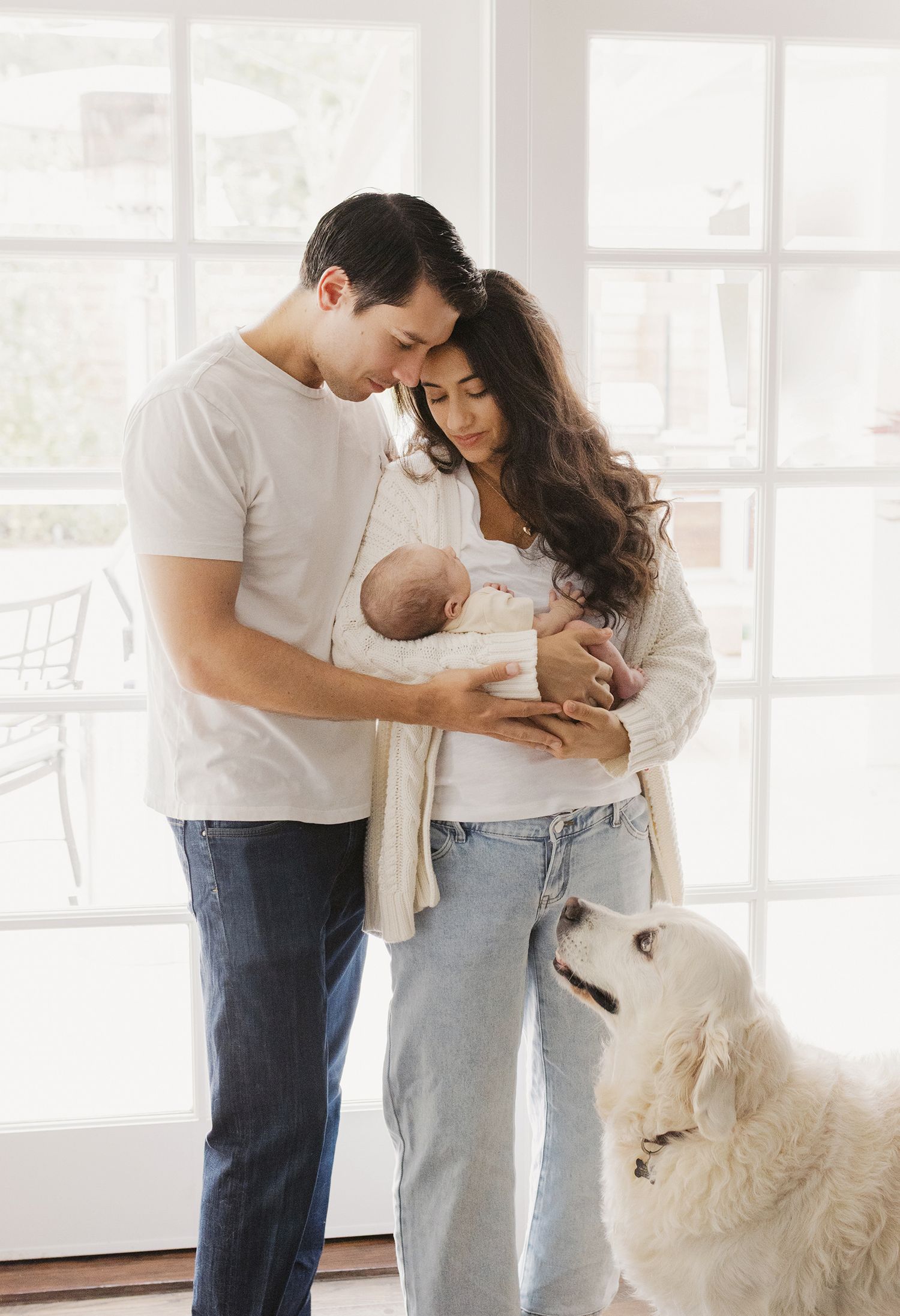 A young couple gazes lovingly at their newborn baby while a fluffy white dog sits nearby in a bright, airy home.